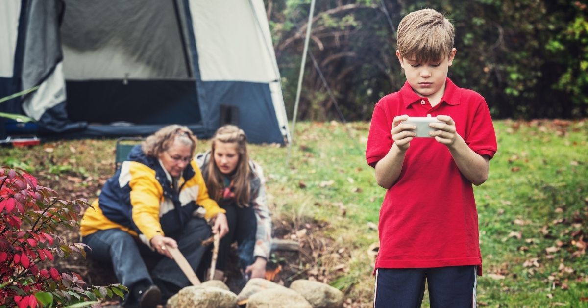 Little boy playing a video game stands apart from mother and daughter who are lighting a fire in front of a tent.